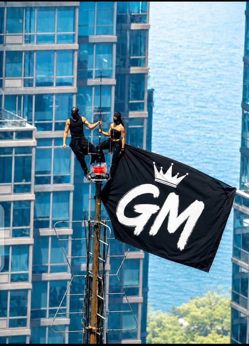 Two masked figures in black outfits perch atop a communications tower, holding a large black flag with a white 'GM' logo and crown, set against a backdrop of tall blue glass buildings.