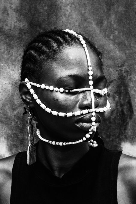 A striking black-and-white portrait of a Black woman adorned with intricate beaded facial jewelry, her eyes closed in serene contemplation. The image complements the post's celebration of West African artistry, embodying the cultural richness and craftsmanship highlighted by the "Ìtàn" NFT collection on objkt.com.