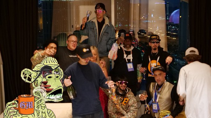 A group of people posing together at an indoor party, holding drinks, with an animated green character holding a "GM" mug overlaid on the image.