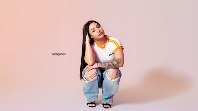 A young woman with long dark hair posing against a pink background, wearing a white and yellow t-shirt and ripped jeans, with tattoos on her arm; text reads '/isafiqueroo'.
