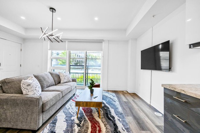 A bright living room features a gray sectional sofa, a wall-mounted TV, a modern coffee table on a patterned rug, and a view through sliding doors to a balcony.