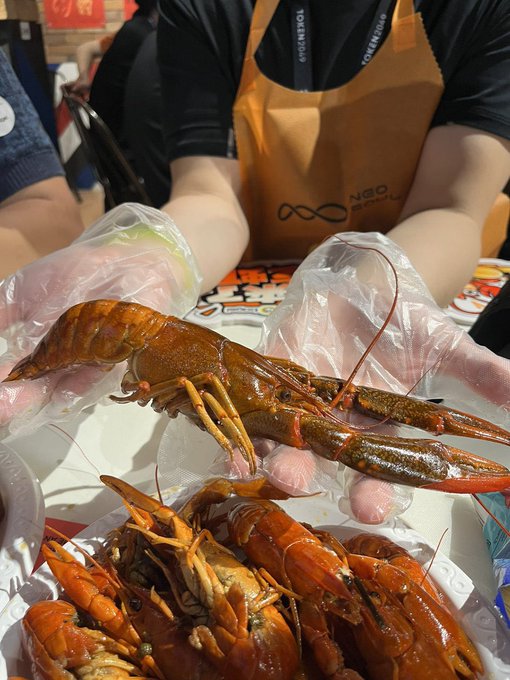 A person wearing plastic gloves holds up a large cooked crawfish, with more crawfish on a tray and a 'NEO GOLF' apron visible in the background.