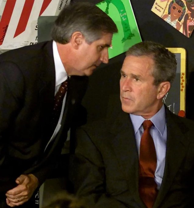 A photograph of two men in suits, one leaning over to whisper to the other who is seated, against a background of children's drawings.