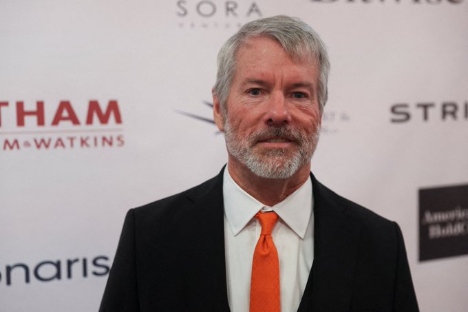 A gray-haired, bearded man in a dark suit, white shirt, and bright orange tie poses on a red carpet in front of a sponsor backdrop featuring logos including "Sora Ventures," "GTHAM McWatkins," and "STR[ipes]." The image does not appear to directly relate to the accompanying post about Strategy (formerly MicroStrategy) selling MSTR shares, as no identifying names, tickers, or numbers matching the financial content are visible.