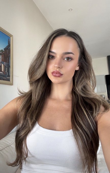A close-up selfie of a young woman with long, wavy brown hair and full lips, wearing a white ribbed tank top, standing indoors with a framed picture of a building visible on the wall behind her.