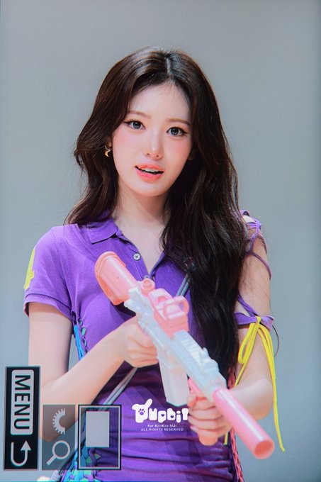 A young woman with long dark hair wearing a purple polo shirt holds a pink and white water gun, appearing to pose for a camera screen displaying a "MENU" and zoom options. The "BugBug" text visible on her shirt, along with the hashtags "#SUI #KiiiKiii #수이 #키키" in the caption, identifies her as Sui, a member of the K-pop group KiiiKiii, photographed on August 23rd.