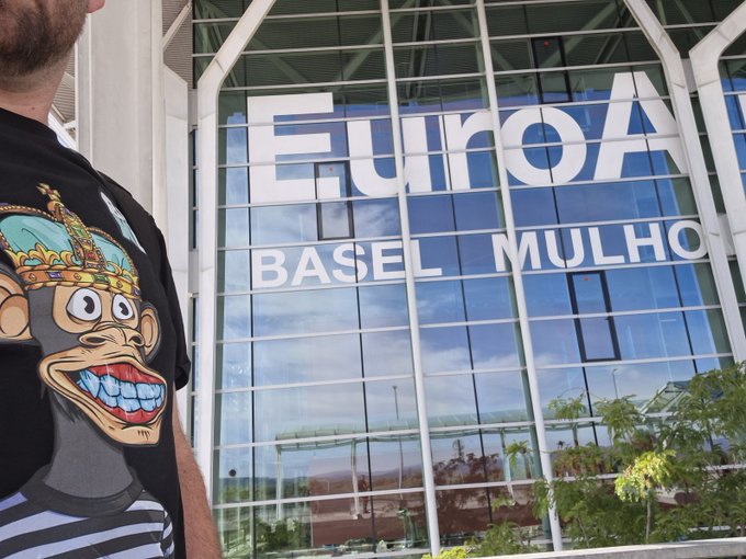 A man wearing a black t-shirt featuring a cartoon monkey wearing a crown stands in front of the EuroAirport Basel Mulhouse Freiburg terminal building with its large glass facade.
