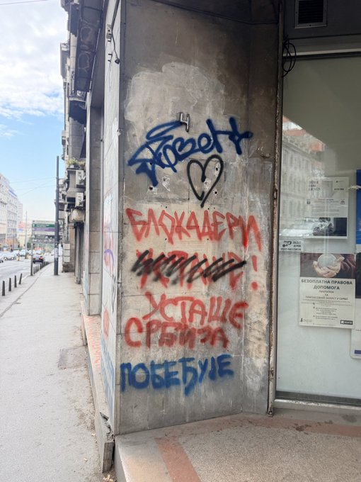 A concrete urban utility box on a sidewalk covered in blue, red, and black graffiti tags, including a heart symbol and Cyrillic text reading 'УСТАЛИЕ СРЫМЗА ПОБЕБУДЕ'.