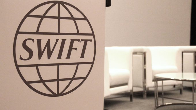 The SWIFT logo is prominently displayed in a corporate meeting room setting. The image relates to the post by showing the branding of SWIFT, the global financial messaging service that HSBC and Standard Chartered used to complete their first real interbank transaction on Swift's new blockchain ledger, with tokenized deposits enabling nearly real-time cross-border payment settlement.