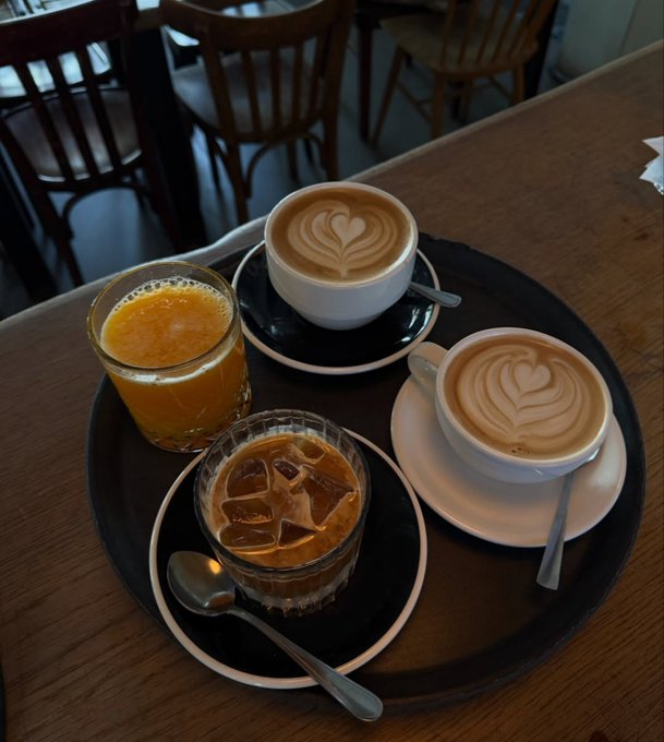 A tray on a wooden table featuring two cappuccinos with heart-shaped latte art, a glass of orange juice, and a glass of iced coffee, with chairs visible in the background.