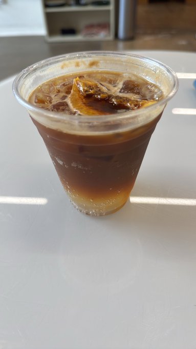 A clear plastic cup of iced coffee with citrus slices and ice, sitting on a white table in a bright room.