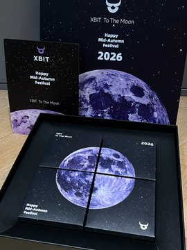 A promotional display for 'XBiT To The Moon' featuring a Happy New Moon Festival 2026 greeting card and a tray of chocolate pieces with a moon design.