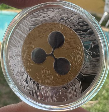 A silver and gold physical cryptocurrency coin featuring the XRP logo on a circuit-board design, held in a protective capsule.