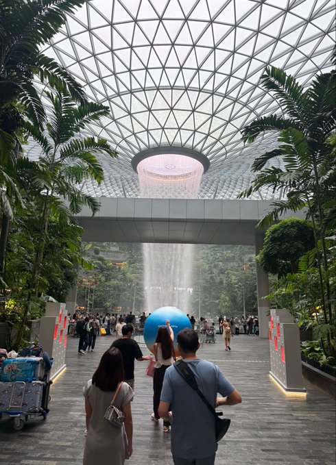 A wide shot inside the Jewel Changi Airport in Singapore, featuring the iconic indoor Rain Vortex waterfall surrounded by lush greenery under a geometric glass dome roof, with visitors walking through the space including a person carrying a large blue exercise ball.