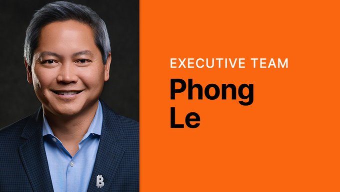 An executive team portrait of Phong Le, wearing a dark blazer over a blue shirt with a Bitcoin logo pin, set against an orange background.