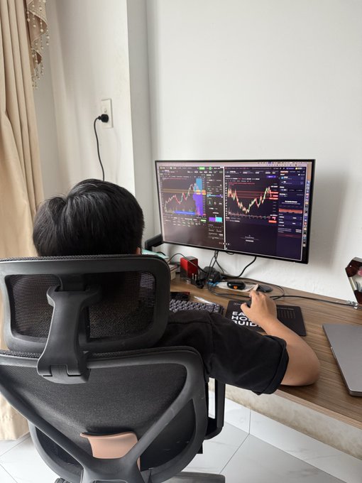 A person sitting in an ergonomic chair at a desk, viewing cryptocurrency charts on a wide monitor.