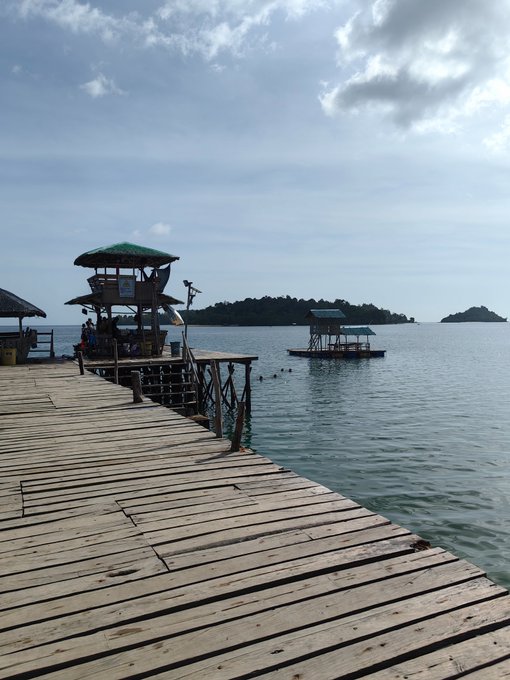 A wooden pier extends over calm ocean water toward a small stilted structure with a green thatched roof, with tropical islands visible in the distance under a partly cloudy sky.