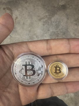 A close-up of a hand holding two physical Bitcoin tokens, one large silver coin and one smaller gold coin, against a gray background.