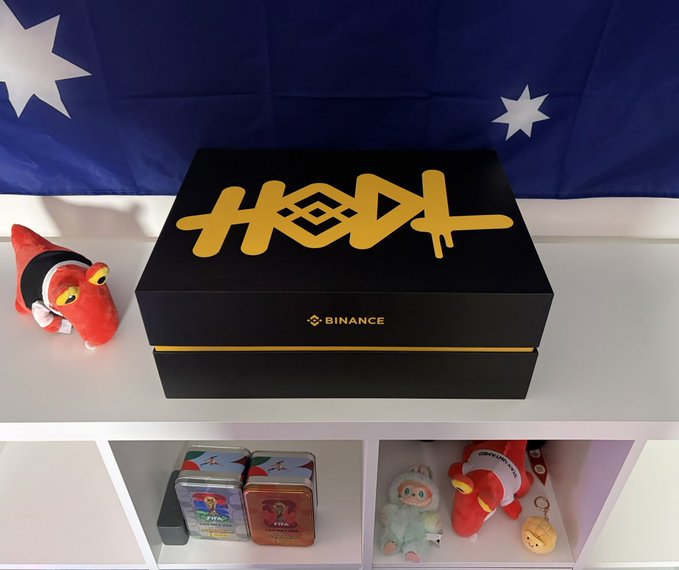 A black Binance box with a yellow graffiti-style logo sits on a shelf against an Australian flag backdrop, surrounded by small plush toys and collectible tins.