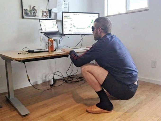 A humorous image of a man in business attire squatting at a standing desk while looking at stock charts on dual monitors, illustrating unusual trading setups.