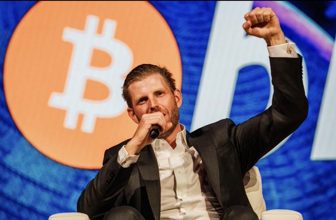 A man in a dark suit holds a microphone and raises his fist on stage in front of a large Bitcoin logo backdrop.