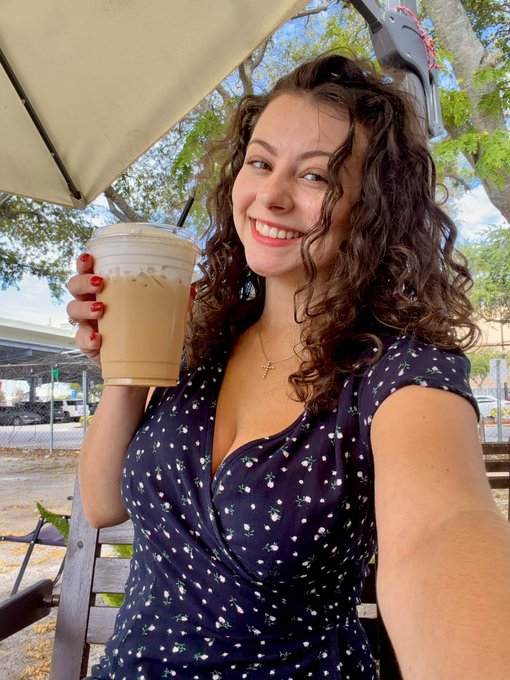 The image features a woman sitting at an outdoor table with a cup of coffee in her hand. She has a smile on her face as she enjoys her drink. The background includes trees, indicating that they are likely outdoors. There is also a bench visible behind the woman and another person seated nearby.