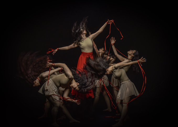 A group of dancers perform a dramatic routine in beige tops and red skirts, captured mid-motion with flying hair against a black background. They hold long red ribbons that flow through the air around them.