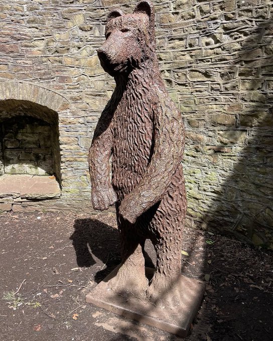 A textured metal sculpture of a standing bear is positioned in front of an old stone wall with an arched alcove.