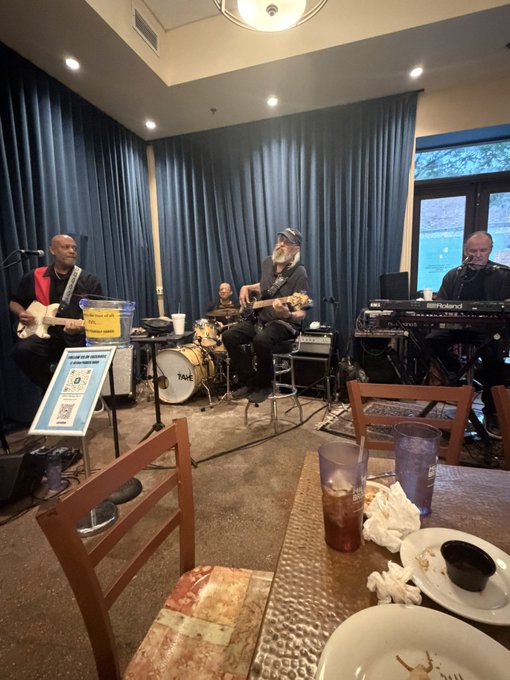 A small jazz trio performs in a lounge, with a drummer seated behind a drum kit labeled "TAKE," a bassist with a red guitar strap, and a keyboardist playing a Roland keyboard. A patron's dining table with iced drinks and empty plates sits in the foreground, capturing the casual "jazzmaxxing" vibe of enjoying live music with dinner.