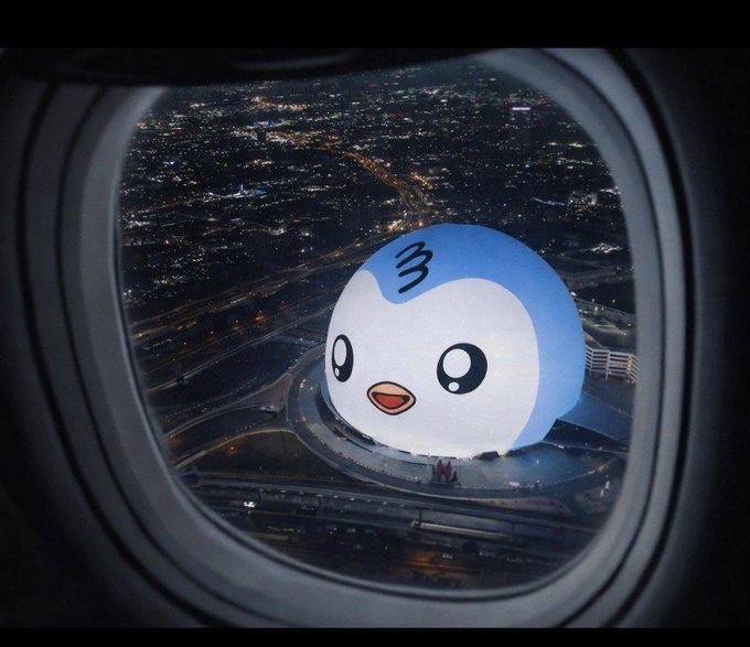 A nighttime view through an airplane window showing a cityscape with highways below, while a large illuminated spherical billboard displaying a cute blue and white penguin mascot is visible on the ground.