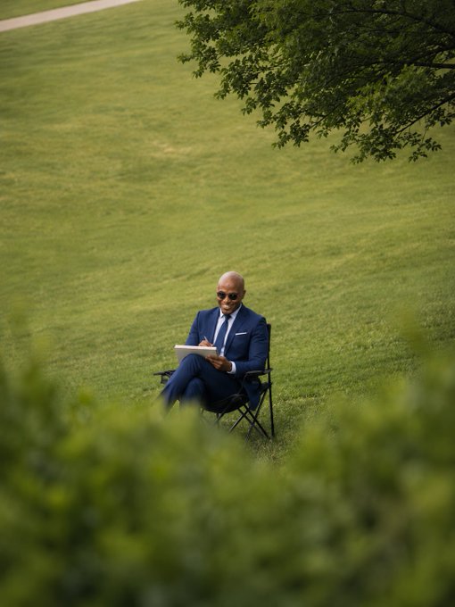 An older man in a blue suit and sunglasses sits in a folding chair on a green lawn, looking down at a tablet, framed by overhanging tree branches.