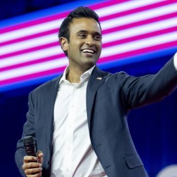 A smiling man in a dark suit with an American flag pin on his lapel, gesturing with his arm raised on a stage with red and blue striped background lighting.