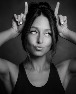 A black and white portrait of a woman making bunny ears with her hands behind her head.