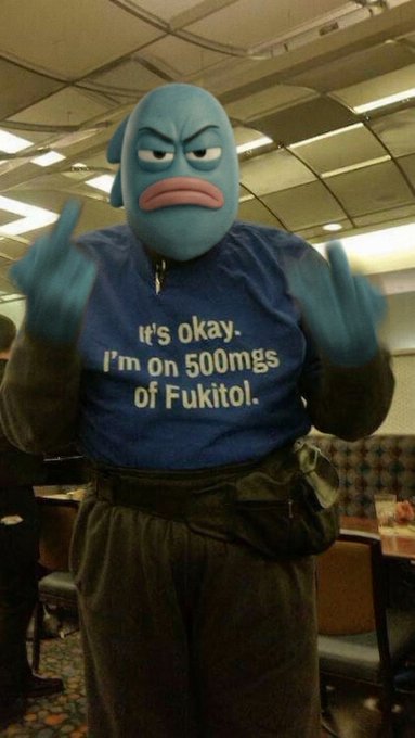 A person in a blue fish-like mask wearing a blue t-shirt that reads "It's okay. I'm on 500mgs of Fukitol." with both middle fingers raised.