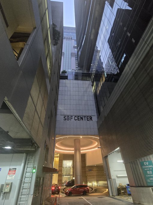 A nighttime photograph of the entrance to SBF CENTER, a high-rise building, with an EV charging station visible at the bottom right and a red car parked under the building's illuminated rotunda.