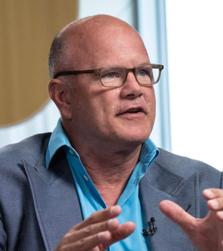 A bald man wearing glasses, a blue shirt, and a grey blazer gestures with his hands while speaking, appearing to be in an interview setting.