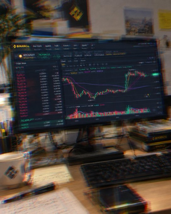 A blurred image shows a computer monitor displaying the Binance trading platform with the BTC/USDT pair at 76,494.01, accompanied by a keyboard, notebook, and coffee mug on a cluttered desk—visually capturing the overwhelming, eye-straining experience of staring at charts all day.