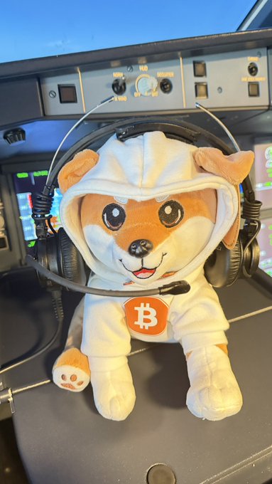 A plush Shiba Inu dog wearing a hoodie with a Bitcoin symbol and aviation headphones sits in an aircraft cockpit.