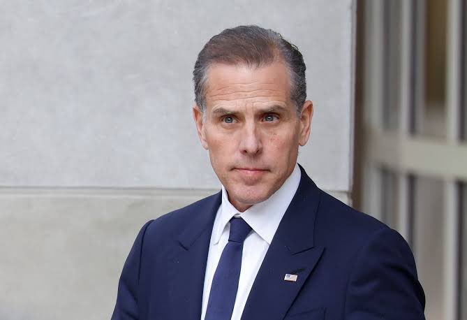 A man in a dark blue suit, white shirt, and navy tie stands outside a building, wearing a small American flag lapel pin.
