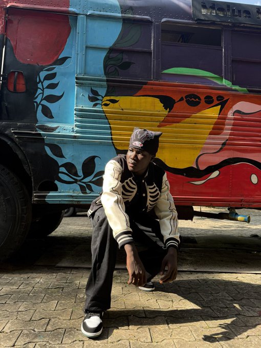 A young man wearing a cap and varsity jacket crouches in front of a vibrantly painted bus with floral and face designs.