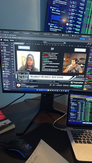 A trading setup displays a monitor showing a YouTube live stream titled "Market Bubble: Sep 24th" featuring hosts including "Banks," alongside another screen filled with cryptocurrency market data and tickers. A laptop and keyboard sit on the desk below, with handwritten notes and a mouse nearby—capturing a trader's workspace while engaging with the tagged accounts @MarketBubble, @Banks, @rasmr_eth, and @phantom.