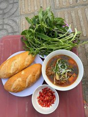 A meal of Vietnamese soup (likely bún bò Huế) served with fresh herbs, lime, red chili, and two baguettes on a red table.