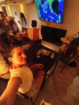 A woman takes a selfie in a lively room where several people are gathered, with a TV screen showing a sports broadcast in the background.