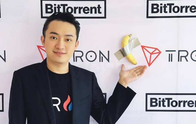 A man in a black blazer and TRON t-shirt posing with a banana taped to a wall, with 'BitTorrent' and 'TRON' logos visible in the background.