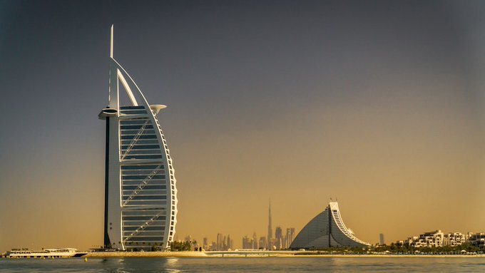 The Burj Al Arab hotel in Dubai stands prominently against a hazy orange sky, with the Dubai skyline and another distinctive sail-shaped building visible in the background across the water.