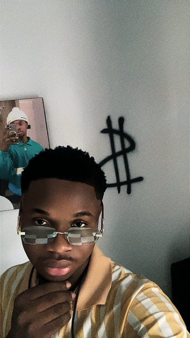 A man wearing rimless glasses poses thoughtfully in front of a wall featuring a spray-painted dollar sign ($) and a poster, matching the "Gm mutuals" vibe with his casual, confident morning selfie aesthetic.