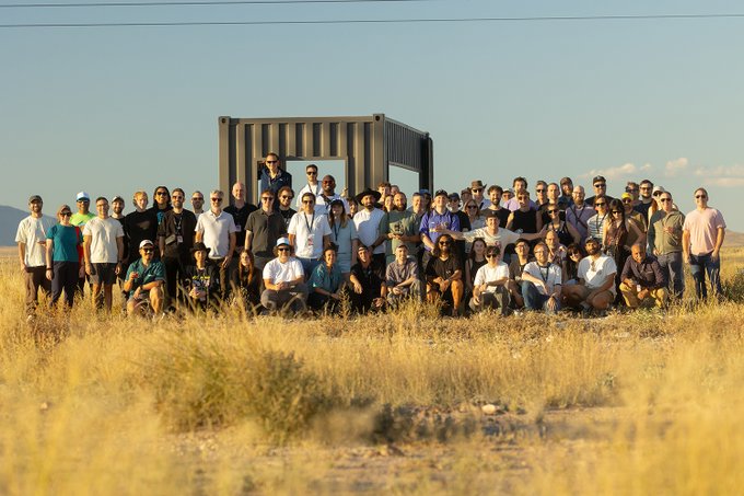 A group of around 40-50 people pose for a photo in a dry grassy field in front of one of Donald Judd's iconic "100 untitled works in mill aluminum" concrete box sculptures, gathering for what appears to be an art and generative art field trip.