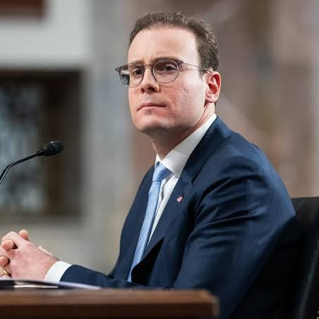 A man in a navy blue suit, white shirt, and light blue tie sits at a desk with a microphone, appearing to be at a formal hearing or testimony. His composed expression relates to the post announcing a regulatory shift away from targeting Bitcoin software developers.