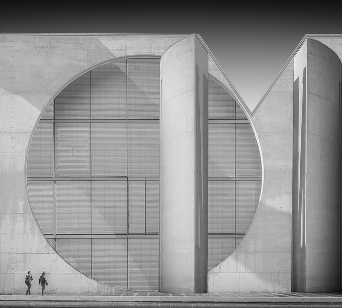 A black-and-white architectural photo of a massive concrete building facade featuring a large circular window and the letter "M," with two small figures walking at the base.