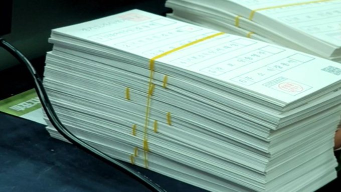 A thick stack of papers or documents is bundled together with yellow rubber bands, featuring printed tables and a red stamp on top, resting on a surface with a black cable.